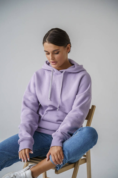 Portrait of beautiful young brunette in purple hoody and blue jeans posing on gray studio background - Photo, image