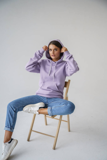 Portrait of beautiful young brunette in purple hoody and blue jeans posing on gray studio background - Fotó, kép