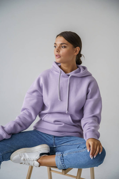 Portrait of beautiful young brunette in purple hoody and blue jeans posing on gray studio background - Фото, изображение