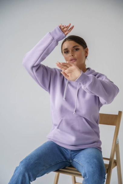 Portrait of beautiful young brunette in purple hoody and blue jeans posing on gray studio background - Foto, Imagem