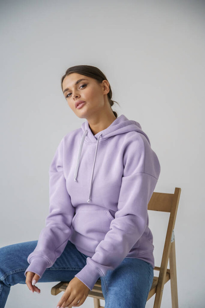 Portrait of beautiful young brunette in purple hoody and blue jeans posing on gray studio background - Photo, image
