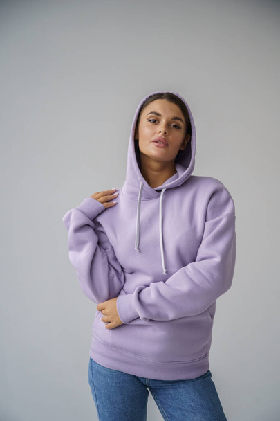 Portrait of beautiful young brunette in purple hoody and blue jeans posing on gray studio background - Foto, imagen