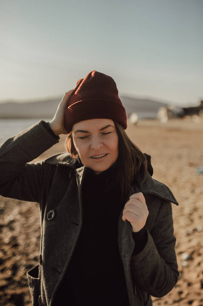 ungefiltertes ästhetisches Porträt eines jungen Mädchens in vielschichtiger Kleidung, mit kastanienbraunem Hut, grauem Mantel auf dem Hintergrund eines Sandstrandes am Meer, selektiver Fokus - Foto, Bild