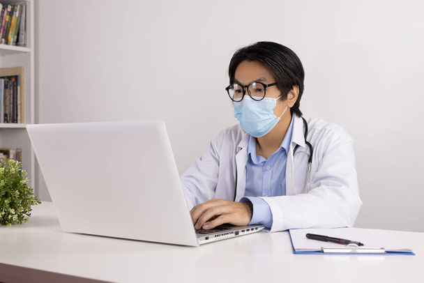 Giovane asiatico medico uomo in laboratorio cappotto o abito con stetoscopio indossare maschera facciale e occhiali utilizzando computer portatile sul tavolo medico in ufficio - Foto, immagini