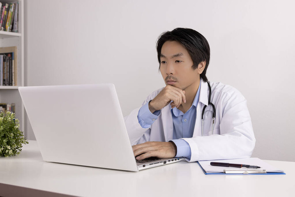 Ernsthafter junger asiatischer Arzt im Laborkittel oder -kleid mit Stethoskop-Touchkinn und Laptop-Bedienung auf dem Arzttisch im Büro - Foto, Bild