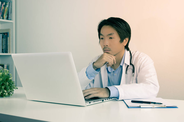 Serious Young Asian Doctor Man in Lab Coat or Gown with Stethoscope Touch Chin and Using Laptop Computer on Doctor Table in Office in Vintage Tone - Photo, Image