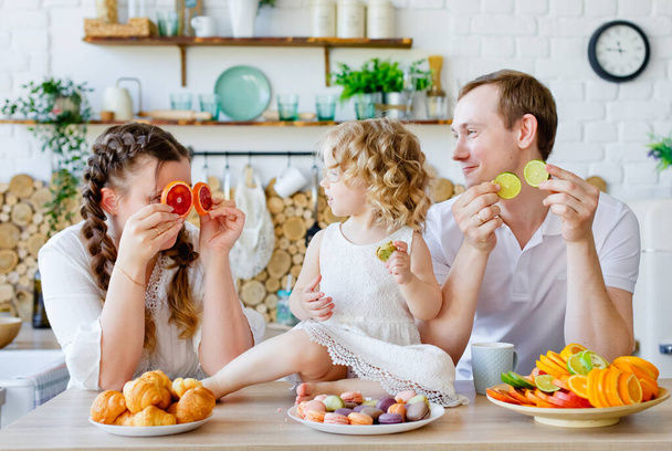 Perhekuva onnellisesta äidistä, tyttärestä ja isästä, jotka poseeraavat keittiössä aamiaisen aikana, syövät herkullisia makaroneja, kakkuja, keksejä. Naimisissa hyvät suhteet, rakastakaa toisianne. - Valokuva, kuva
