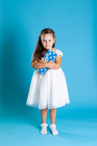 full-length little Princess in white dress and snow petal makeup clutches Christmas gift to her chest, isolated on a blue background - 写真・画像