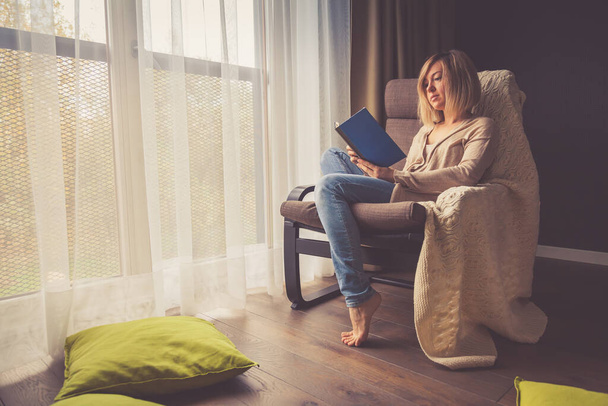 Молода жінка ізолюється вдома і розслабляється, вона читає книгу, що сидить у кріслі біля вікна
 - Фото, зображення