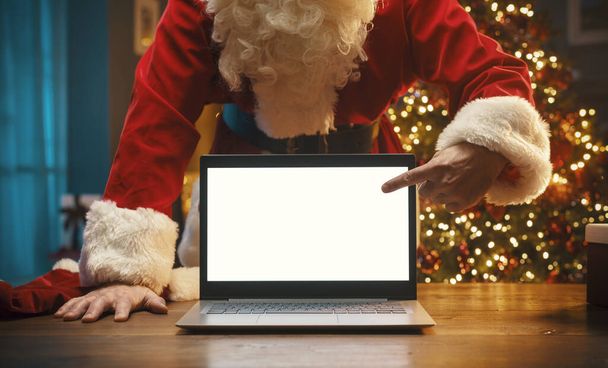 Santa Claus standing behind a desk and pointing at the laptop screen, Christmas and communication concept - Valokuva, kuva