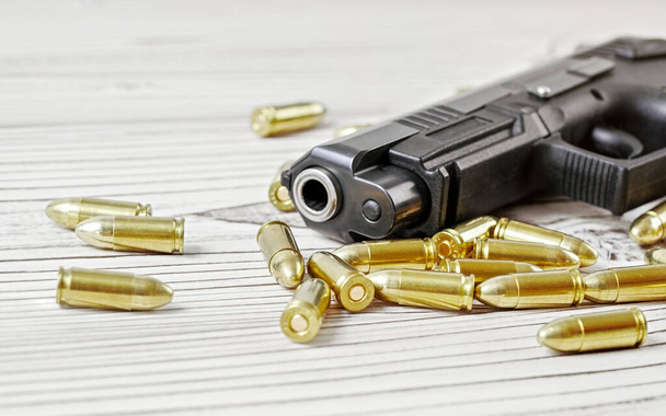 Closeup of modern dark gray gun with yellow brass bullets next to it on white wooden boards - Foto, immagini