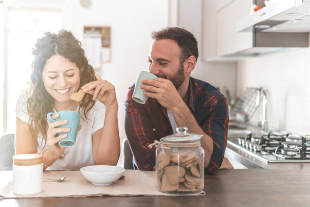 Engaged couple has breakfast together in their new home - young couple smiling while drinking and eating in the kitchen - warm filter on background - Photo, Image