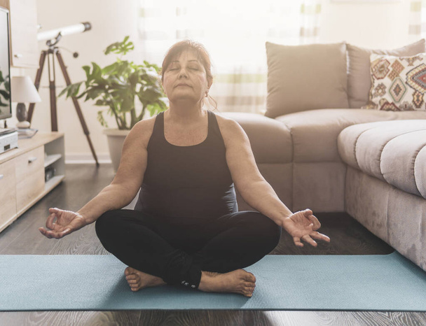 Middle aged woman sitting in yoga position with closed eyes - Senior woman relaxes meditating in her home - Healthy life and wellness concept - warm filter on background - Photo, Image