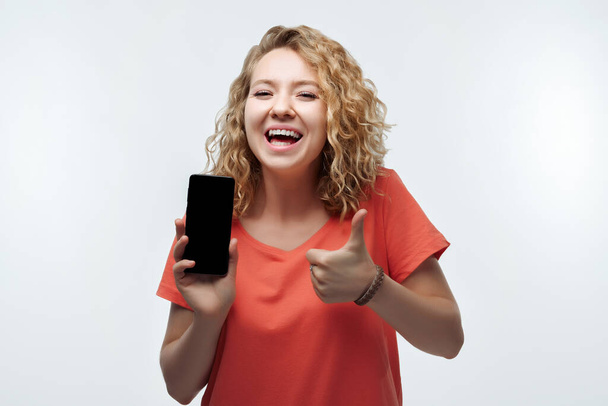Молодая женщина с кудрявой прической, одета в повседневную футболку Показывает смартфон пустой экран, глядя на камеру Рекомендуем мобильное приложение - Фото, изображение