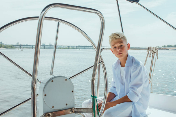 Serious teen handsome blonde boy in white t-shirt and shirt on the yacht. Education, lessons, studying, skills, growing up concept - Photo, Image