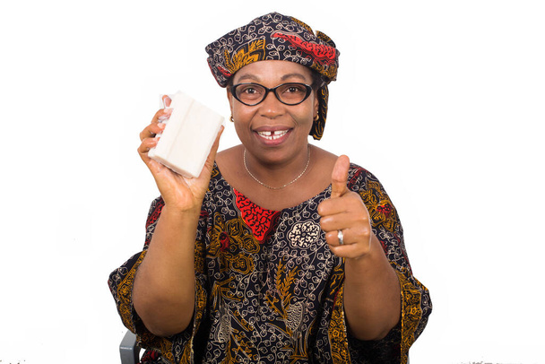 Glückliche Frau in afrikanischem Outfit mit Schal auf dem Kopf hält weiße Seife in die Kamera und zeigt sie mit Handbewegung isoliert auf weißem Hintergrund. - Foto, Bild