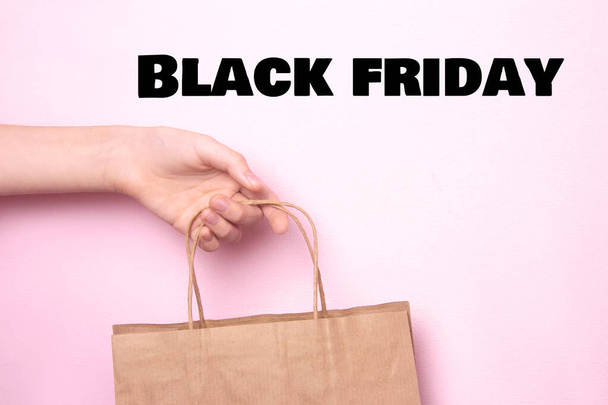 Female hand holds a craft bag for shopping on a bright pink background. Ecological paper shopping bag in the hands of a woman. Black Friday sale. Copy space - 写真・画像