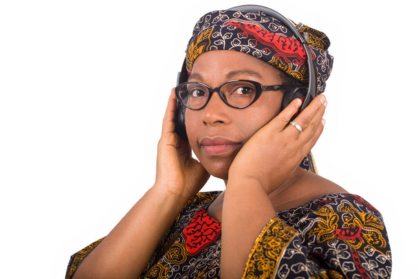 hermosa mujer afroamericana escucha música. Mujer africana en vestido tradicional con una bufanda en la cabeza de perfil y sosteniendo un auricular en la oreja mientras mira a la cámara - Foto, imagen