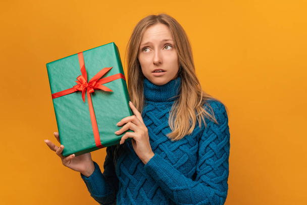 Портрет молодої жінки в в'язаному синьому светрі, що тримає подарунок, прикрашений стрічкою. Студійні зйомки, жовтий фон. Новий рік, жіночий день, день народження, святкова концепція - Фото, зображення