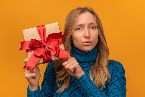 Portrait of a charming young woman in knitted blue sweater holding gift decorated with ribbon. Studio shot, yellow background. New Year, Women's Day, Birthday, Holiday concept - Photo, Image