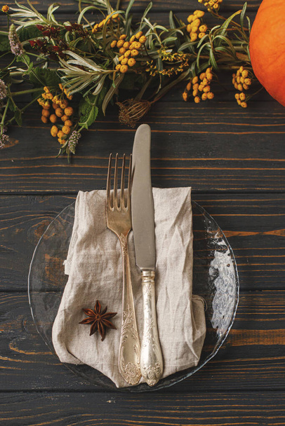 Thanksgiving table setting. Stylish vintage cutlery, linen napkin, anise star on modern glass plate on background of autumnal floral arrangement and pumpkin on rustic wood. Flat lay. Eco friendly - Photo, Image