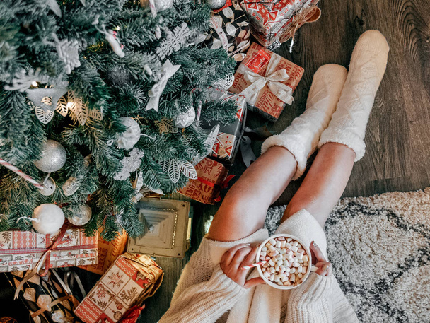 une vue de dessus d'une fille caucasienne portant des chaussettes blanches floues, assise sous le sapin de Noël, et buvant du chocolat chaud avec des mini guimauves - Photo, image