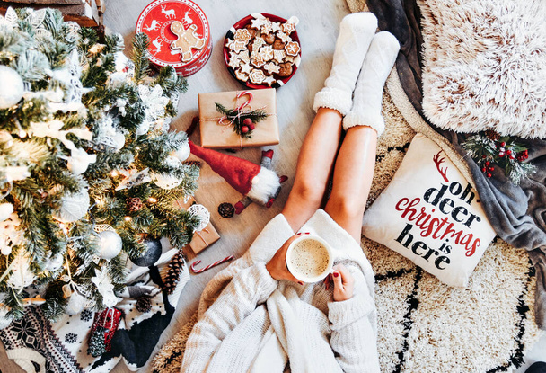 A top view of a caucasian girl wearing white fuzzy socks, sitting under the Christmas tree, and drinking hot chocolate - Fotoğraf, Görsel