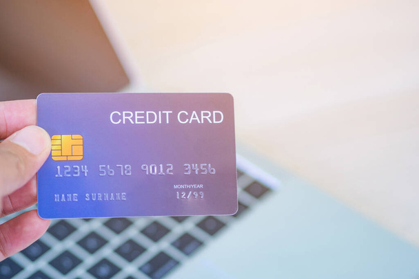 Close-up photo of the left-hand woman holding blue credit card mockup with a laptop background and copy space for text. Easy e-commerce shopping and payment by credit card. Concept of e-commerce - Photo, Image