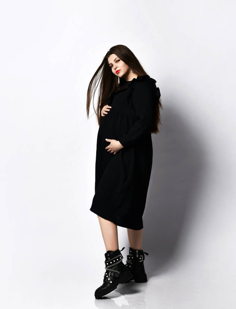 Pregnant brutal woman in long black dress and brutal shoes stands side to camera holding hands on her belly - Photo, Image