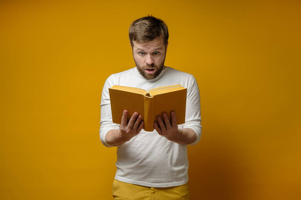 Atrakcyjny młodzieniec z entuzjazmem czyta książkę, stojącą na żółtym tle. Koncepcja edukacji. - Zdjęcie, obraz