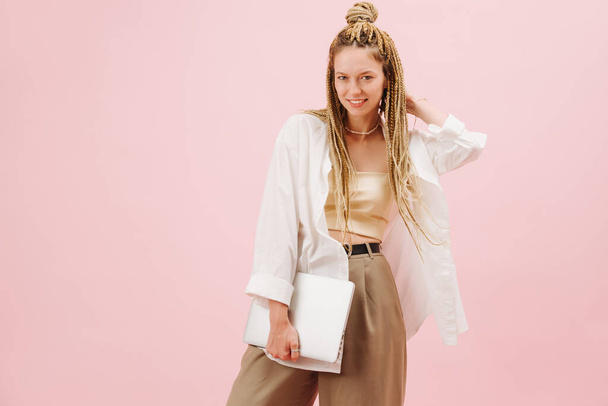 Stunning eccentric young woman with stylish blond afro braids over pink background. She's posing for a photo, one hand on a back of her head, holding notebook in a second hand. - Foto, Imagem