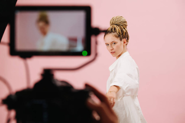 Attractive woman with blond afro braids in a bun posing in front of a camera with a field monito. Over pink background. Blurred equipment in a foreground. - Φωτογραφία, εικόνα