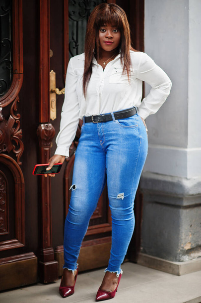 Cute african american model in white shirt and jeans pants posed outdoor with mobile phone at hand. - Фото, изображение