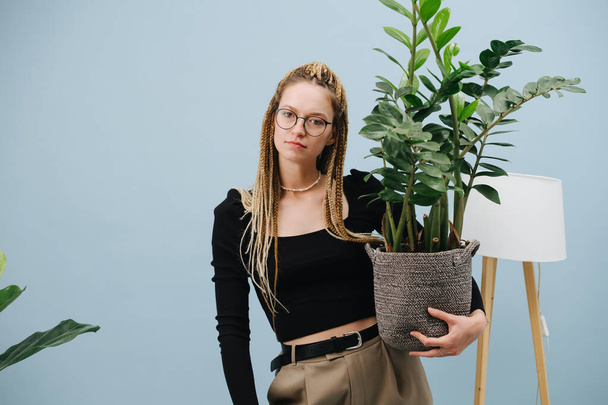 Casual young woman with stylish blond afro braids over blue background. She's posing for a photo with big potted plant in hand. - Fotó, kép
