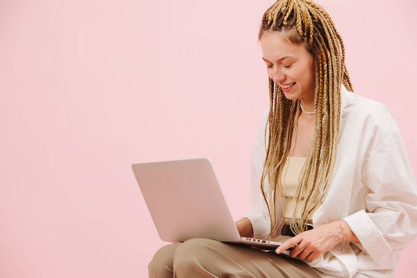 Boldog fiatal nő, elegáns szőke afro zsinórral, aki egy laptopon dolgozik rózsaszín háttérrel. Mosolyog, becsukja a szemét.. - Fotó, kép