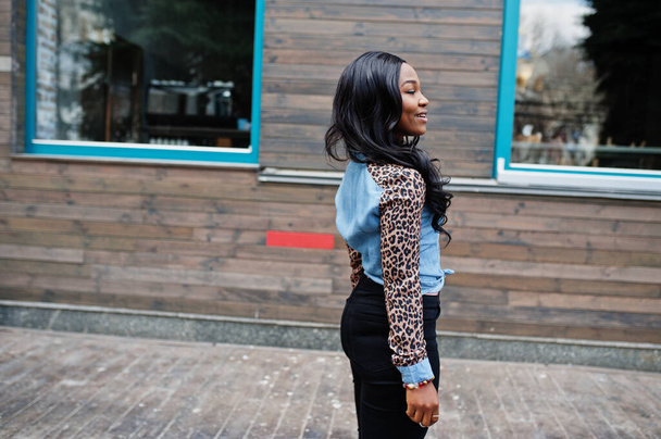 Hipster african american girl wearing jeans shirt with leopard sleeves posing at street against wooden house with windows. - Fotó, kép