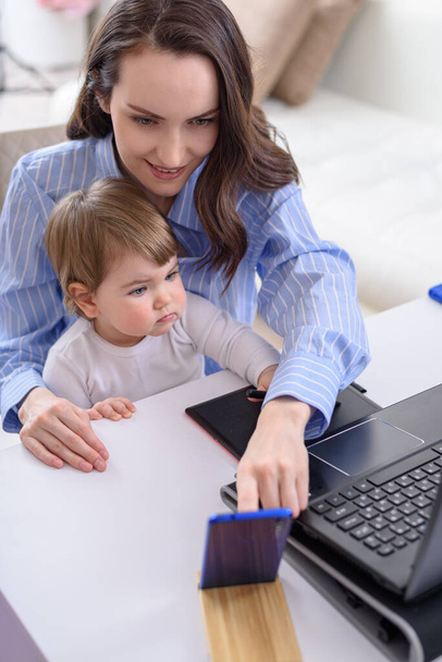 uśmiechnięta brunetka z dzieckiem siedząca przy laptopie, odbierająca rozmowy wideo, zdalna praca w domu z dziećmi, pionowa - Zdjęcie, obraz