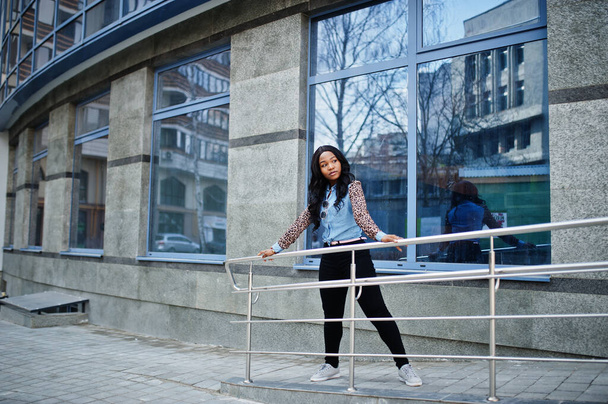 Hipster african american girl wearing jeans shirt with leopard sleeves posing at street against modern office building with blue windows. - Foto, afbeelding