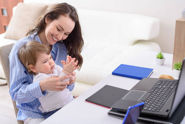 uśmiechnięta kobieta w niebieskiej koszuli z dzieckiem siedzącym na laptopie bawiącym się w palmtopy, szczęśliwa koncepcja macierzyństwa, zdalna praca w domu z dziećmi, - Zdjęcie, obraz