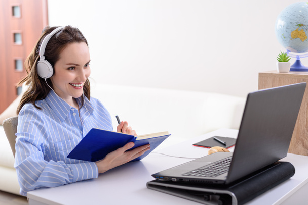 молодая улыбающаяся девушка в синей рубашке с наушниками, сидя за ноутбуком, онлайн-курсы, электронное обучение, удаленная работа, онлайн обучение - Фото, изображение