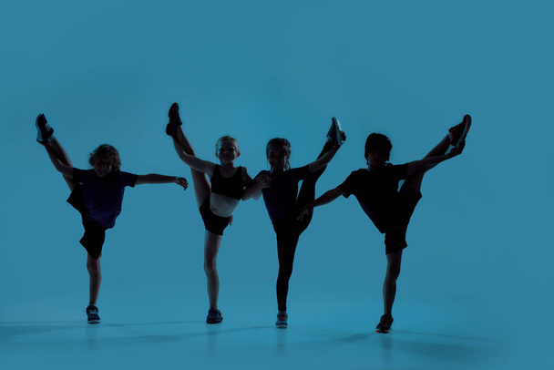 Silhouetted full length shot of four little kids, gymnasts showing flexibility while posing, standing isolated over blue background - Foto, Imagem