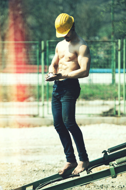 Strong Muscular Man Preparing for Workout in Industrial Junk Yard - Young Athlete Rubs Magnesium on Hands and Wearing Yellow Helmet - Zdjęcie, obraz