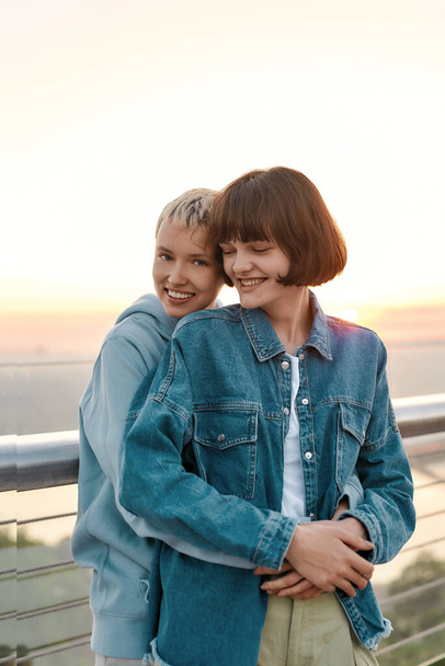 Young loving lesbian couple looking happy, embracing, standing on the bridge, Two women smiling while admiring the sunrise together - Foto, imagen