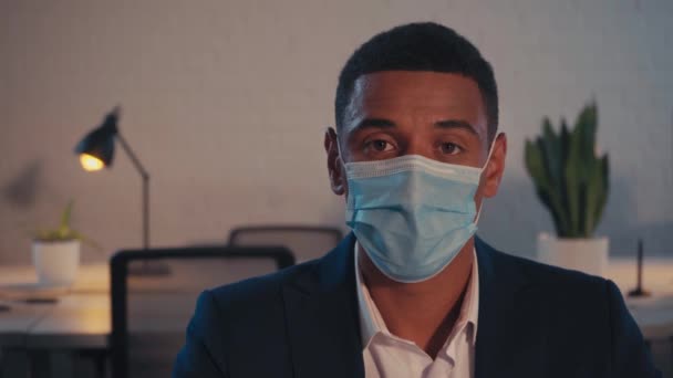 Hombre de negocios afroamericano en máscara médica mirando a la cámara en la oficina  - Imágenes, Vídeo