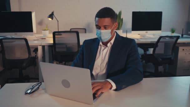 Afrykański amerykański biznesmen w masce medycznej piszący na laptopie w biurze w nocy - Materiał filmowy, wideo