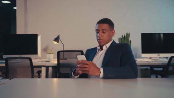 african american businessman messaging on smartphone in office at night - Záběry, video