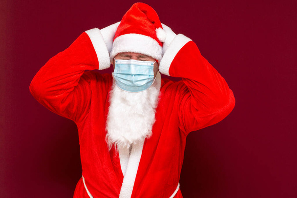 Primer plano Santa Claus mantener la cabeza cerrada ojos máscara médica haciendo un gesto de parada con las manos en el fondo rojo - Foto, Imagen
