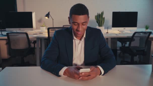 glimlachende Afrikaanse Amerikaanse zakenman chatten op smartphone 's nachts in het kantoor - Video