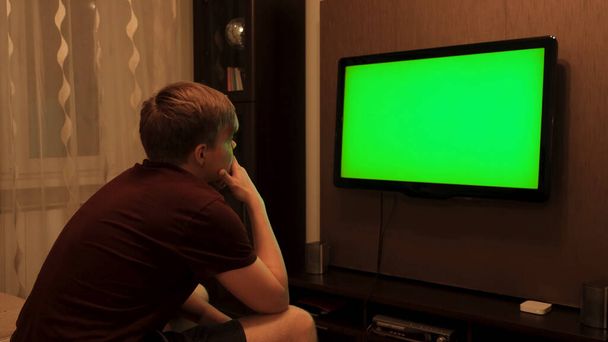 Vue arrière de l'homme regardant la télévision verte. Concept. Homme regarde la télévision avec écran vert et appuie sur la télécommande. Homme change de chaînes à la télévision avec écran vert - Photo, image