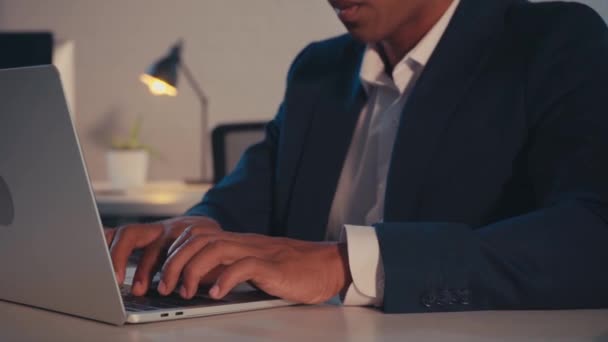 przycięty widok afrykańskiego amerykańskiego biznesmena piszącego na laptopie w nocy w biurze - Materiał filmowy, wideo